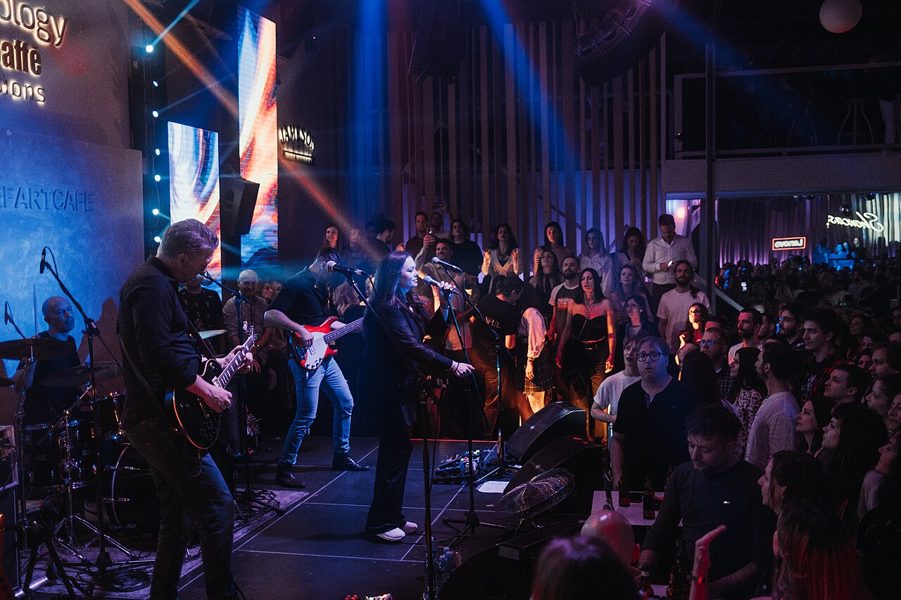 Musician playing guitar on a dimly lit stage with stage lights behind them, audience faintly visible.